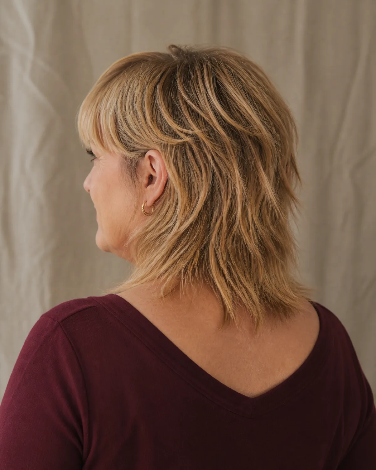 A woman over 50 with a fuller build seen from the back-three-quarter angle, dark blonde shoulder-length shag with choppy disconnected layers at multiple lengths, a heavy fringe partially visible, burgundy V-neck top, linen drape background