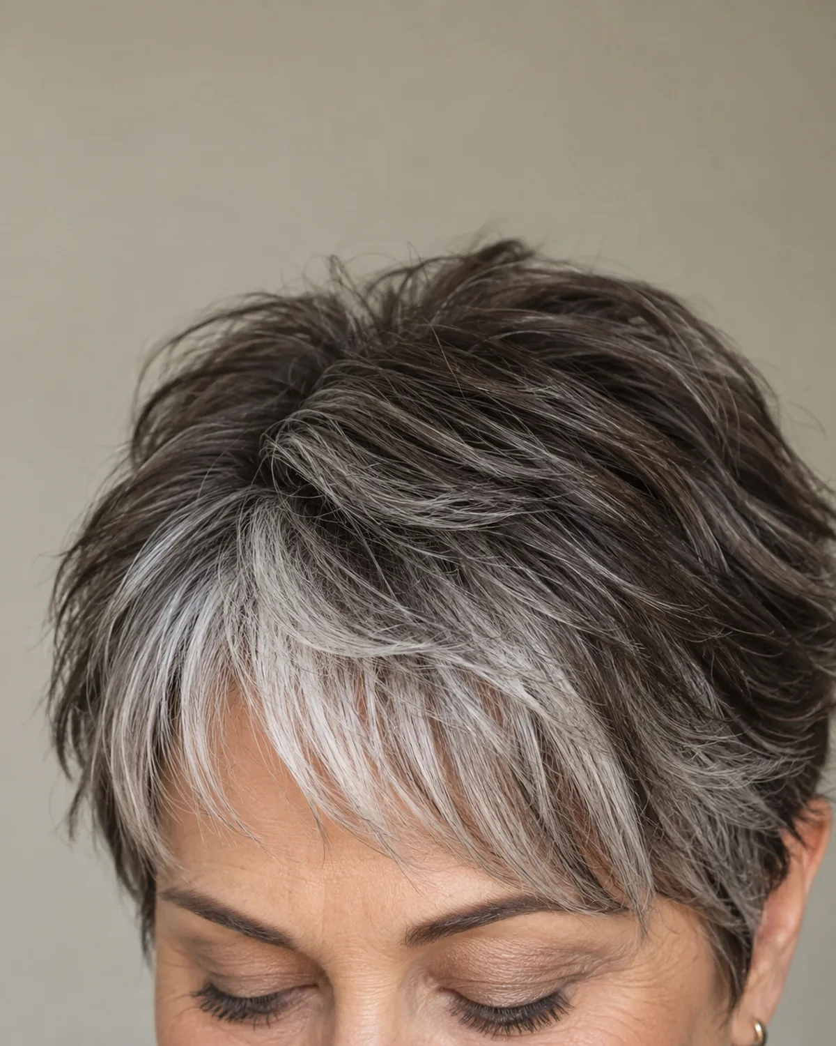 Top-down forehead macro of mature woman with layered crop and silver fringe sweeping across the forehead against darker crown