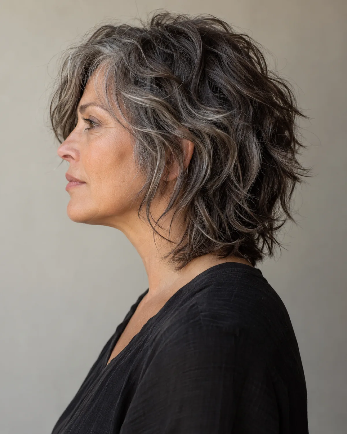 Full left profile of mature woman with textured shag hairstyle, choppy layers showing scattered silver strands through dark hair