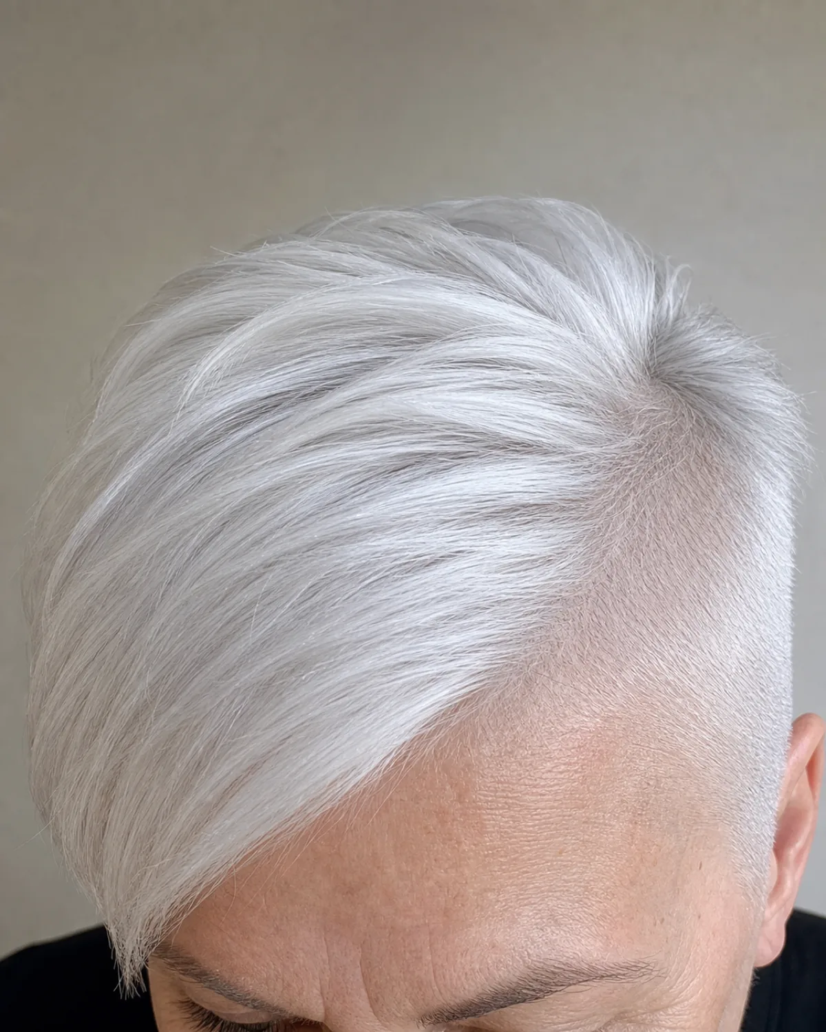 Close crop on the fringe and brow area of a woman in her early 60s with an edgy undercut pixie on bright white hair, long top swept to one side contrasting with a very close buzz at the temple, black crew-neck top