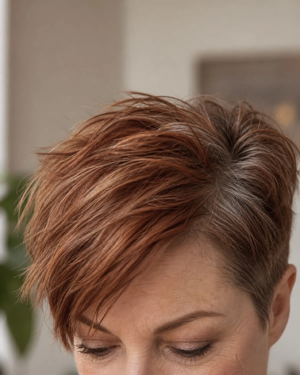 Close crop on the fringe and brow line of a mature woman with thick auburn hair with grey roots in a disconnected pixie, showing long textured top and dramatically short sides