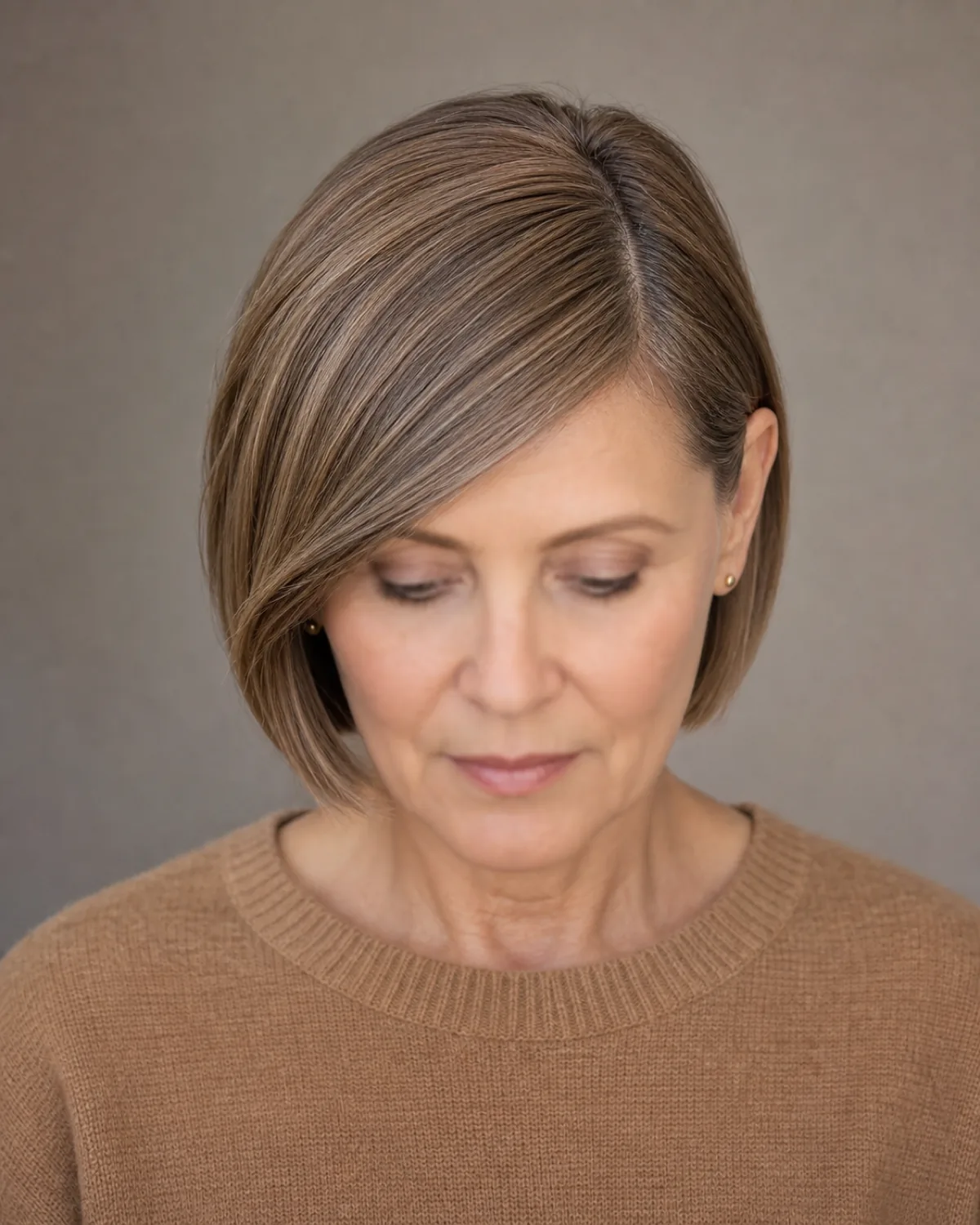 A woman in a camel knit with her eyes lowered, a chin skimming bob with a deep side parting and a fringe sweeping diagonally across her forehead and tucked behind the far ear