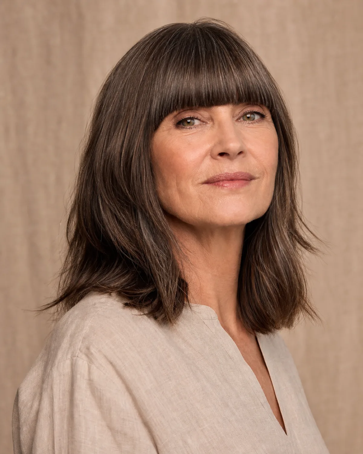 A woman in her late 50s with dark brown shoulder-length hair and a full blunt fringe cut straight across her brow, three-quarter turn with chin slightly lifted, silver strands through the fringe, warm taupe background