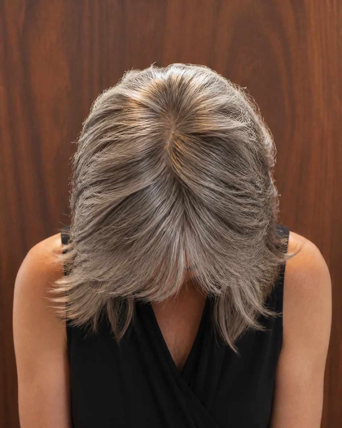 The top of a woman's head tilted forward toward the camera, showing salt-and-pepper shag layers starting short at the crown and graduating longer toward the shoulders, warm walnut panelling behind
