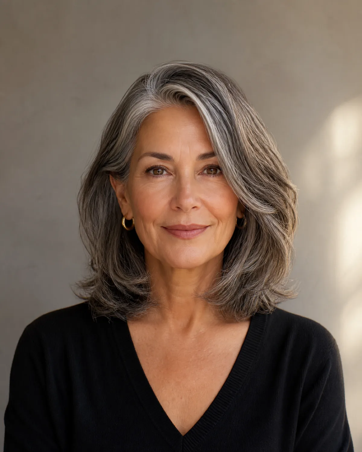 A woman in her early 60s with warm salt-and-pepper hair at shoulder length, a deep side part creating visible asymmetry with more volume on the heavy side, front-facing with features in soft focus