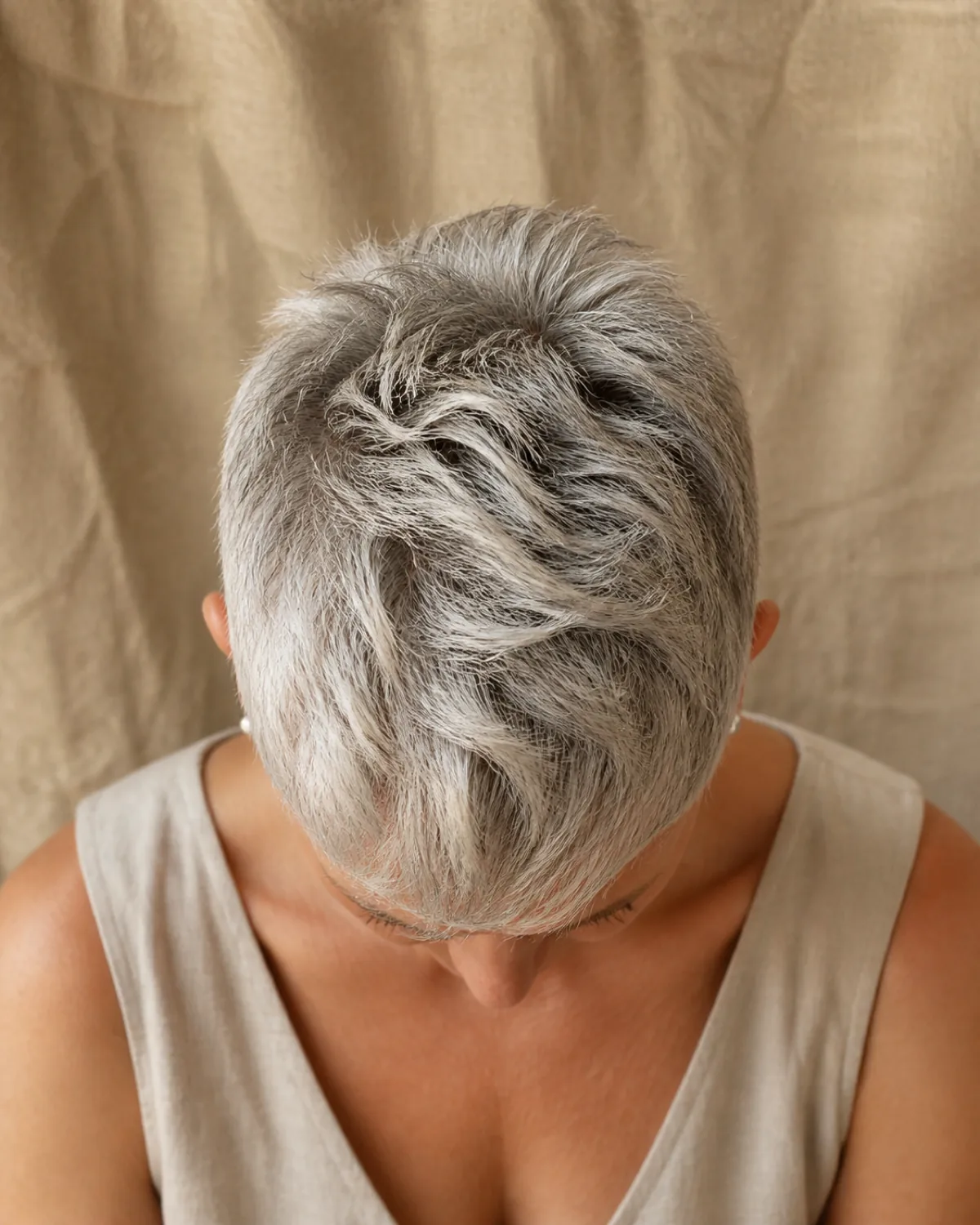 A woman in her late 50s with silver-grey hair in a textured pixie cut, head tilted forward showing the crown, the crown carrying visibly more length and volume than the close-cropped sides