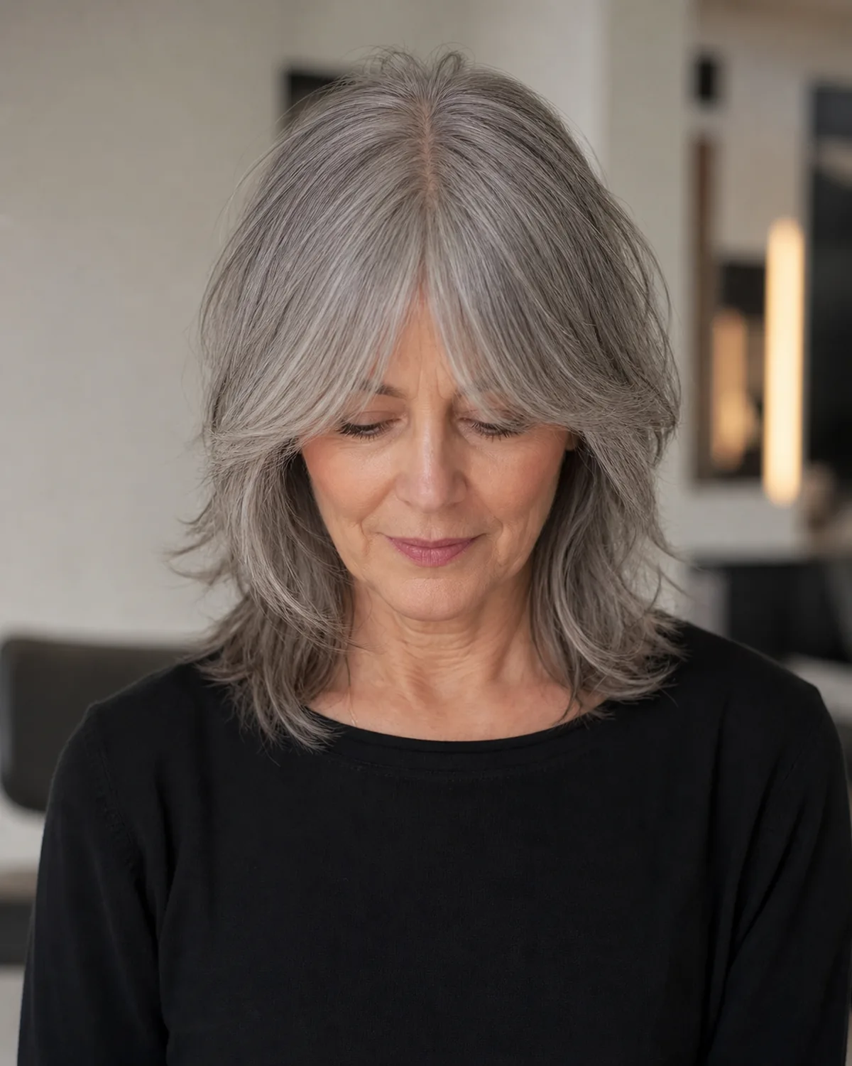 Front-facing view of a mature woman with natural grey hair looking downward, a curtain fringe parting at the center and covering the temple area, with disconnected shaggy layers from the ears down