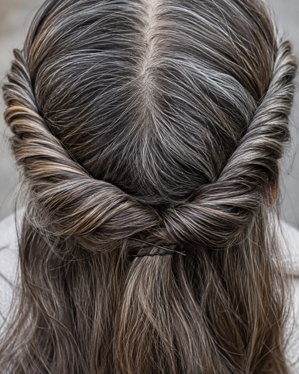 Top-down close crop of a mature woman's crown showing two twisted sections of salt-and-pepper hair converging from each temple, bobby pin at the meeting point, loose hair falling below