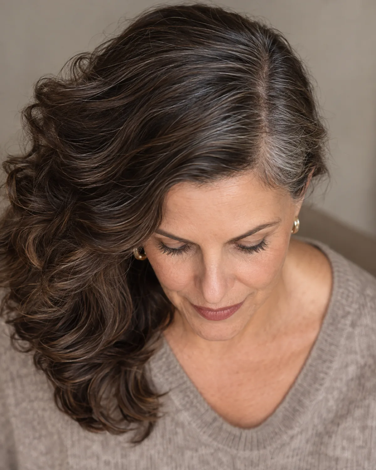 Top-down view of woman over 50 with dark brown wavy hair and silver strands at the part line, deep side part creating volume asymmetry, taupe top