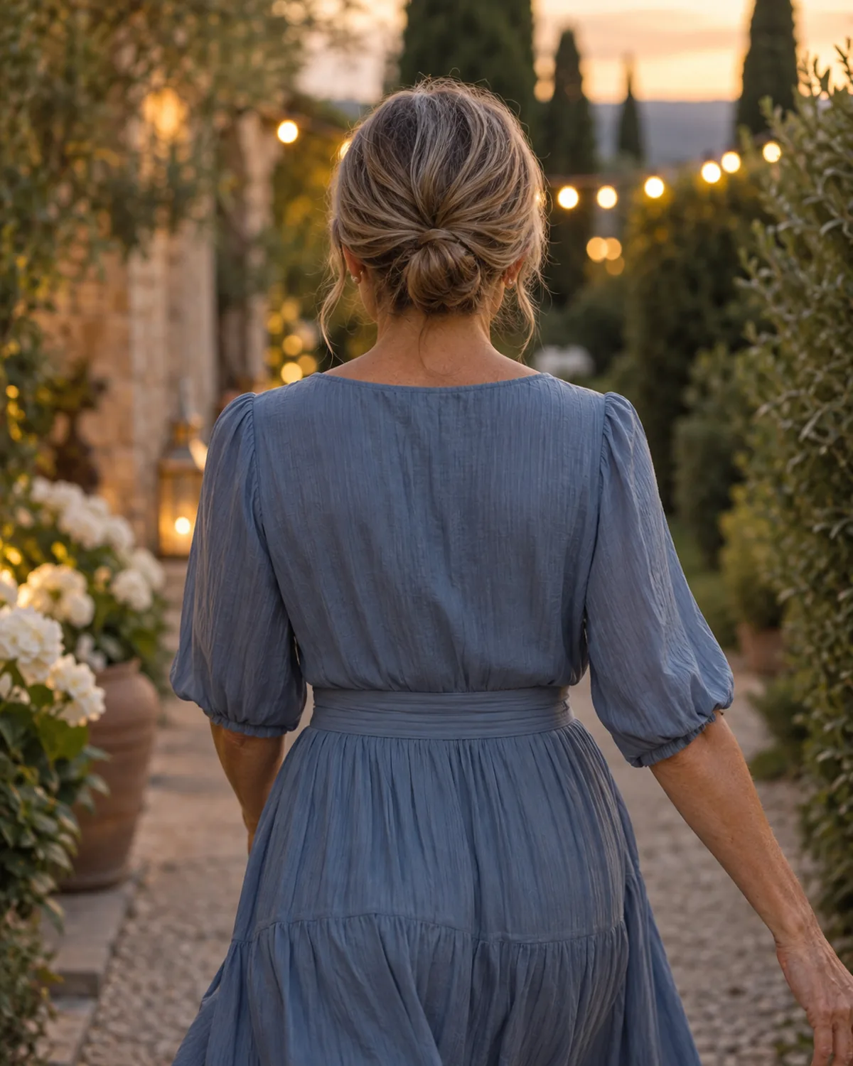 Woman over 50 in a dusty-blue dress walking away down a garden path with fairy lights, her hair in a loose low bun with three pieces pulled out